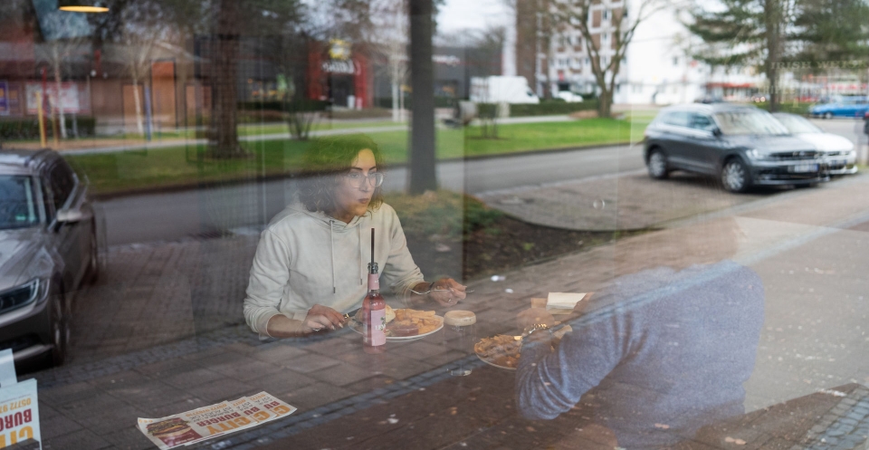 Glückliche Frau isst einen Burger bei City Burger Espelkamp mit Kartoffelspalten und Kaltgetränk, Aufnahme durch die Scheibe von außen